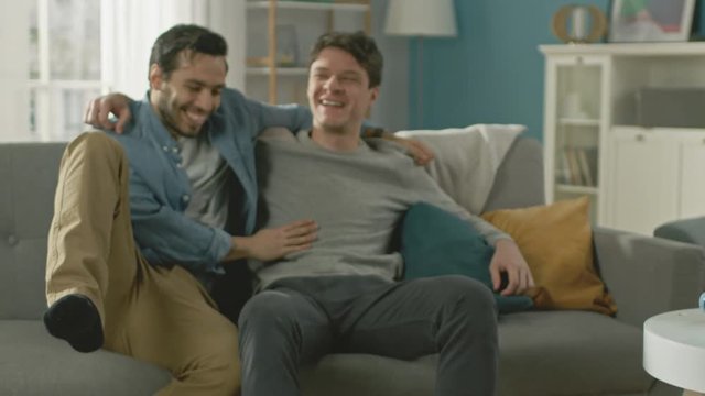 Slow Motion of a Male Gay Couple Fulling Around on a Sofa at Home. Boyfriend Runs and Jumps into Hands of His Partner. They are Happy Laughing. They are Casually Dressed and Room Has Modern Interior.
