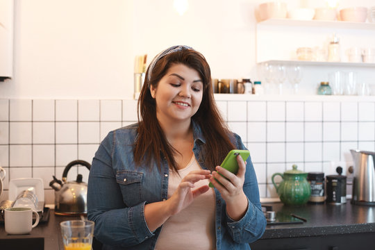 Cheerful beautiful young brunette overweight woman browsing social media on her mobile phone, chatting with friends and leaving comment under nice pic while drinking fresh orange juice in kitchen