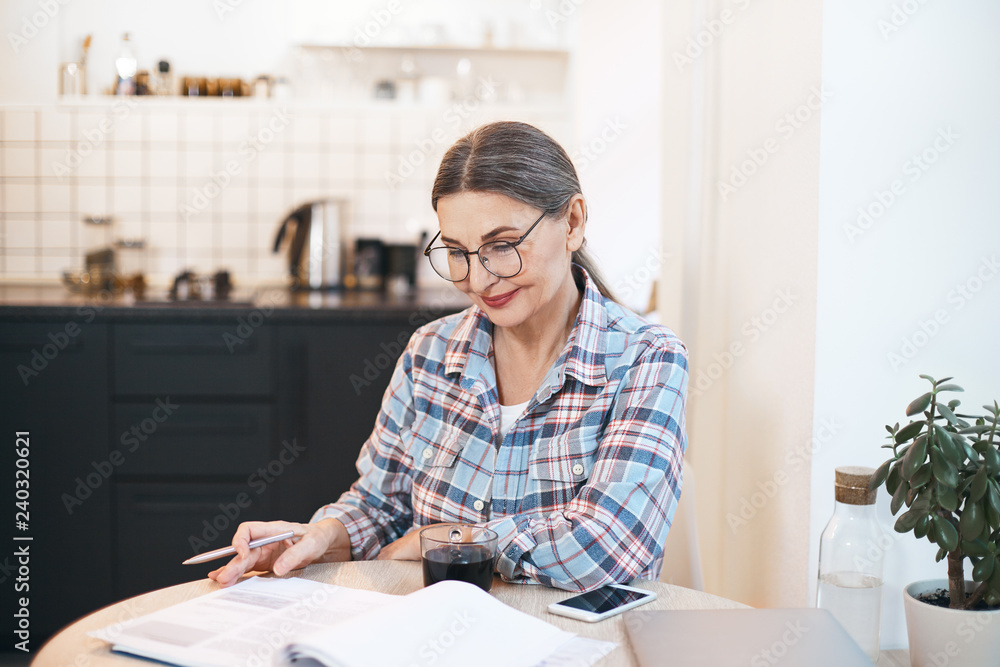 Wall mural People, lifestyle, age, education, paperwork and technology concept. Picture of beautiful positive elderly woman teacher in glasses checking students' copybooks at kitchen table and smiling