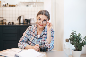 Obraz na płótnie Canvas Cheerful attractive middle aged senior female enjoying tea at kitchen table, touching stylish eyeglasses and smiling broadly, papers and laptop in front of her. People, age and modern technology