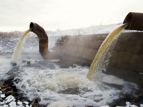 Wastewater From Two Large Rusty Pipes Merge Into The River In Clouds Of Steam