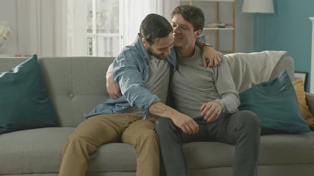 Cute Attractive Male Gay Couple Sit Together on a Sofa at Home. Boyfriend Puts His Hand on Partner's and They Hug. They are Happy and Smiling. They are Casually Dressed and Room Has Modern Interior.