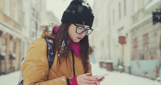 Woman in trendy outfit walks in the winter city passing by local cafes, texts via her phone, browsing the internet. Using online map, gps.