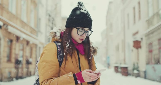 Young woman in trendy outfit walks in the winter city passing by local cafes, texts via her phone, browsing the internet. Using online map, gps.