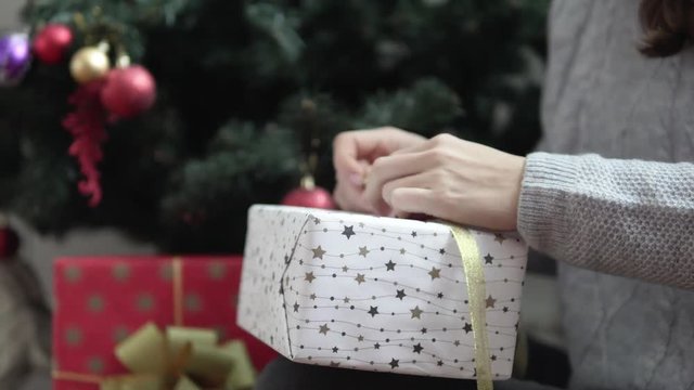 women's hands tied a ribbon on a New Year's gift. The girl is sitting at the Christmas tree, on her lap is a beautiful box, she is tying a golden ribbon. The final decoration of the gift. In the frame