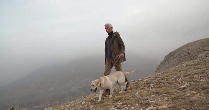 Moustached caucasian pensioner walking with his beautiful golden retriever dog in fogged mountains - old friendship concept 4k