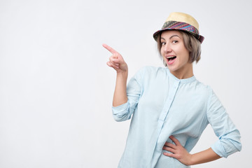 Young attractive woman in summer hat and blue shirt pointing with her finger on copyspace and smiling
