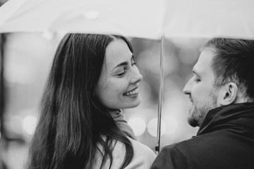 Fototapeta premium Happy beautiful couple, guy and his girlfriend dressed in casual clothes stand under the umbrella and look at each other on the street in the rain. Black and white photo