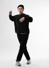 Young freestyle dancer dressed in black jeans, sweatshirt, hat and gray sneakers is dancing in the studio on the white background