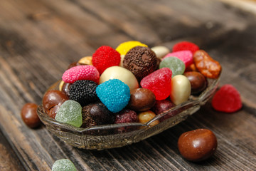 Different candies in a crystal bowl on old wooden table. Multi colored gummy candy coated with sugar, chocolate and fruit candy.
