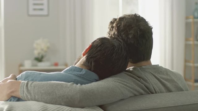 Back Shot of Cute Male Queer Couple Hug While Sitting on a Sofa and Putting Their Heads Together. They are Casually Dressed in Jeans and Sweater. Room has Modern Interior and It's Bright and Sunny.