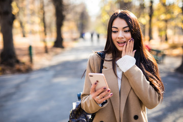 Beautiful young surprised woman read message on mobile phone dressed in autumn coat outdoors