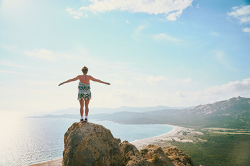 Happy tourist girl standing on the rock high above the big empty sand beach in Corsica