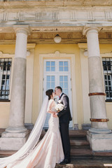 Gorgeous bride in amazing gown and stylish groom kissing and embracing in sunny street at old building. Beautiful happy wedding couple gently hugging. Romantic sensual moment