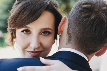 Gorgeous bride and stylish groom gently hugging and embracing in sunny street. Portraits of beautiful happy wedding couple. Romantic sensual moment. True love