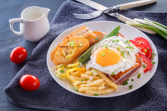 Fried egg with bacon in a white plate with fried pieces of bread, greens, tomatoes and french fries on a gray wooden table. Close-up