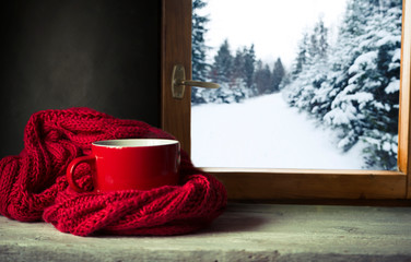 Winter background - cup with candy cane, woolen scarf and gloves on windowsill and winter scene outdoors. Still life with concept of spending winter time at cozy home with cold weather outdoors