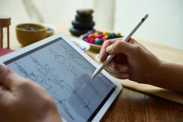 Fashion designer using modern tablet and drawing human figure sketches while working at table in studio