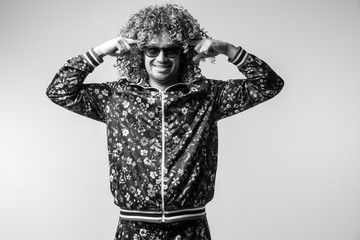 Excited cheerful bizarre man posing om white studio background. Stylish funky male with curly hair black and white portrait. Funny guy in tracksuit expressing feelings. Unusual person in sunglasses.