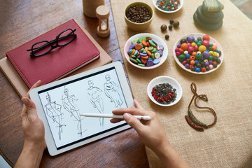 Close-up of female hands sitting at the table with plates with colorful beads and books and drawing sketches of female clothes on digital tablet
