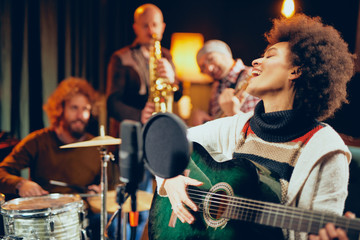 Mixed race woman singing and playing guitar while sitting on chair.In background drummer, saxophonist and bass guitarist. - obrazy, fototapety, plakaty