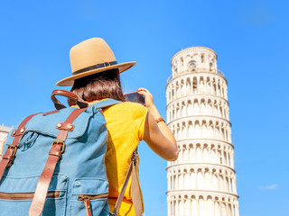 Happy female traveler in hat with backpack taking photo of the leaning tower in Pisa. Vacation and...