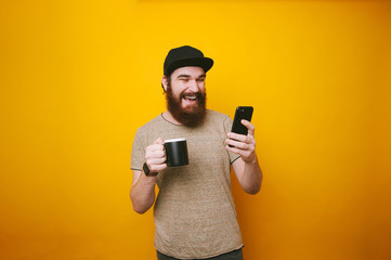 Happy smiling young man is texting on mobile, and holding a cup of tea or coffee