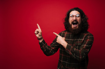 Amazed bar man in shirt and glasses pointing at copy space over red background