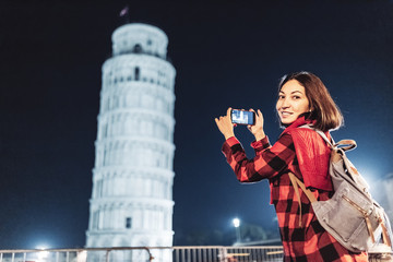 Portrait of happy young woman tourist traveler in front of leaning tower of Pisa, Italy at evening...