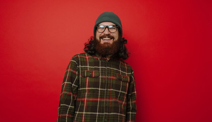 Hipster bearded man in hat and glasses, smiling, standing over red background