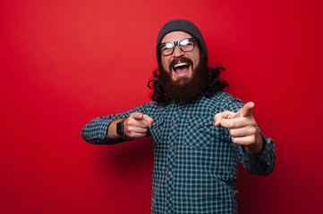 Excited bearded man in hat and glasses is pointing to camera over red background