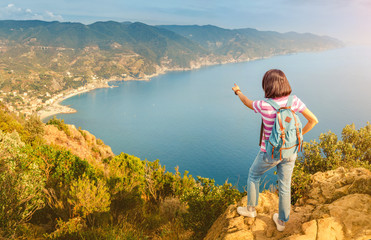 Naklejka premium Happy young woman with backpack enjoying beautiful seascape in Italy. Vacation and travel in Cinqueterre national park, mediterranean sea.
