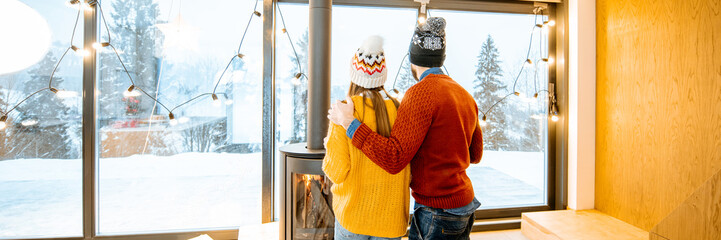 Young couple dressed in bright sweaters enjoying landscape view on the mountains standing together in the modern house during winter time