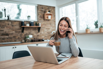 Happy young woman at home, talking on mobile phone and using laptop, drinking hot chocolate.
