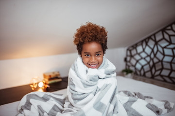 Portrait of a smiling little mixed-race girl with blanket sitting on bed and looking at camera.