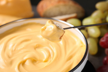 Piece of bread over pot with delicious cheese fondue, closeup