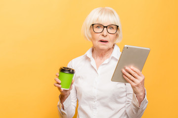 Happy senior woman using tablet while drinking coffee isolated over yellow background.