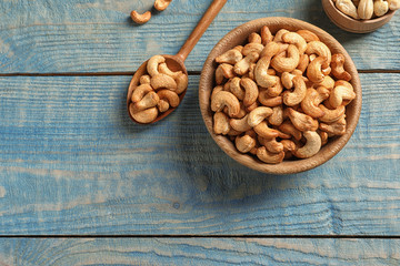 Tasty cashew nuts in bowl and spoon on color wooden table, flat lay. Space for text