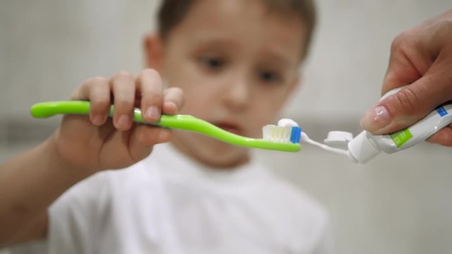 Close-up of the face of a little boy who holds a toothbrush and brushes his teeth. Mom's hand puts white toothpaste on the brush and gives it to the child.