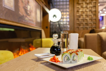 Closeup shogun sushi roll with avocado, strawberry, teriyaki sauce, sesame, wasabi, ginger, white Japanese plate, soy sauce, white napkins, glass water on the tables of lobby of hotel restaurant