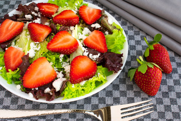 Vegetarian delicious fresh salad with purple and green lettuce leaves, strawberries and goat cheese on grey tablecloth.