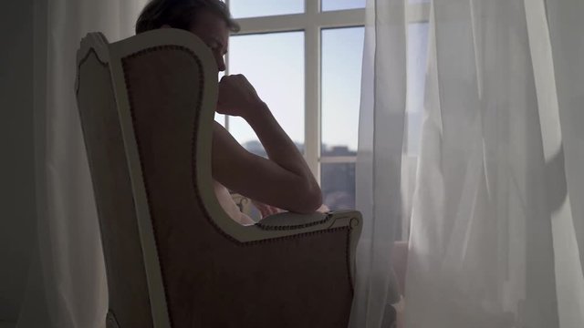 Seriouse young man sitting on the armchair by the window looks into the distance and thinks. Guy with a naked torso reflects on his affairs and makes plans for the future.