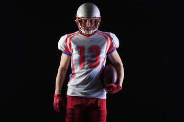 American football player wearing helmet posing with ball on black background