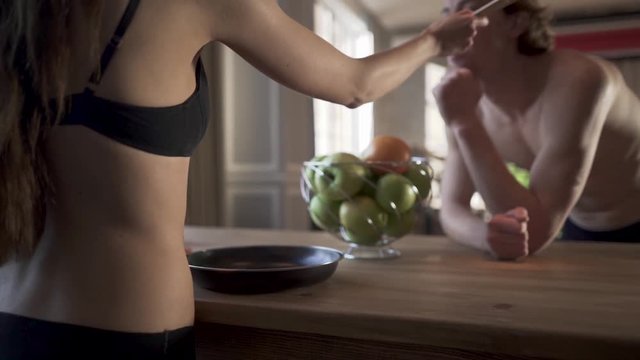 Young naked couple in the loft kitchen. Girl in lingerie give empty spoon for her man to try to eat and man act like its tasty.