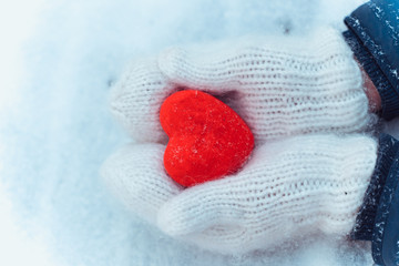 Fototapeta premium Female hands in knitted white mittens with heart of snow in winter day. Love and St. Valentine cozy concept, copyspace