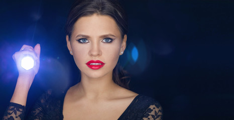 Fototapeta premium girl with the led flashlight in her hands shines straight. . glare of light and dark Studio background. portrait of a beautiful model who looks carefully in the direction of light from the lamp. space