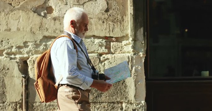 Caucasian old man walking as a tourist in the historic town with a map in hands and looking for the route. Outside.
