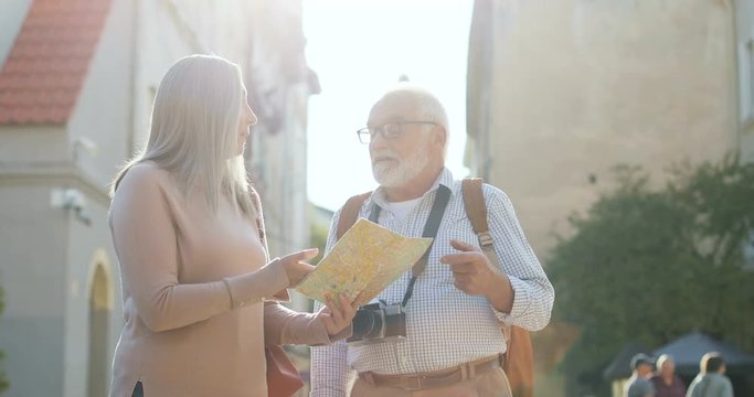 Senior Caucasian couple of tourists standing in the sunlight in the town with a map in hands and looking for the route. Outdoor.