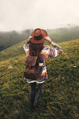  Travel and wanderlust concept. Stylish hipster girl in hat walking on top of mountains. Happy young woman with backpack exploring misty mountains. Amazing atmospheric moment