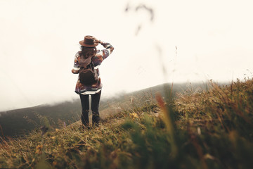 Stylish hipster girl in hat walking on top of mountains. Happy young woman with backpack exploring...